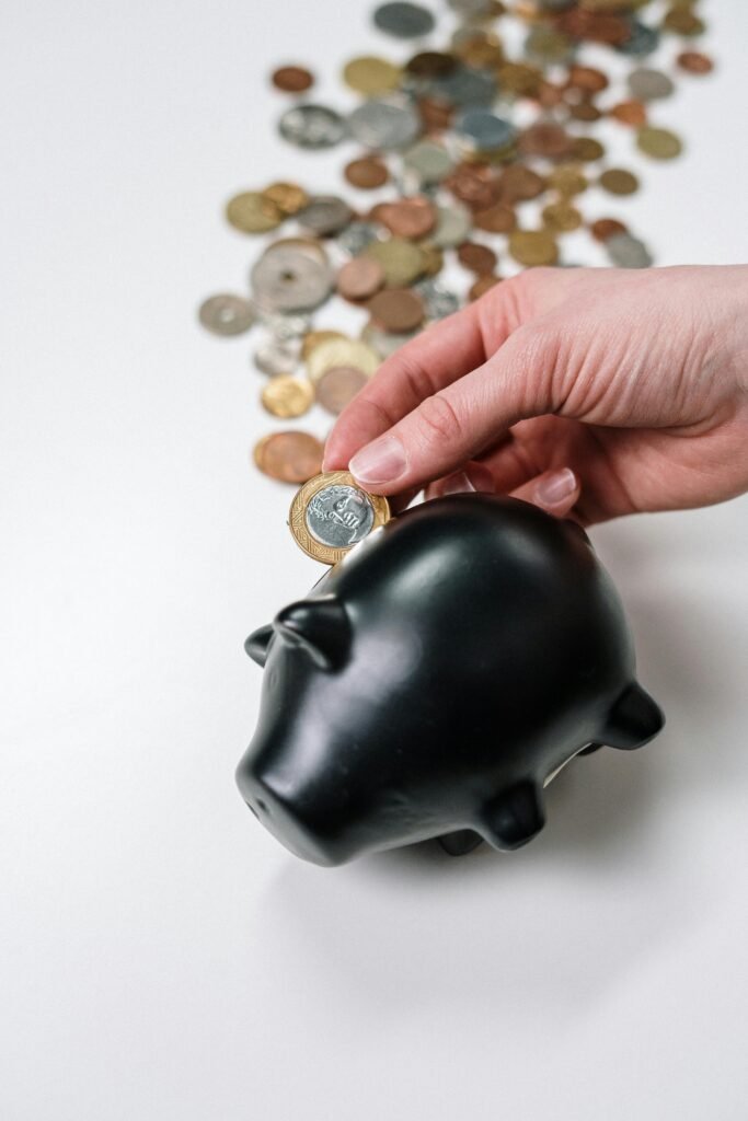 A hand putting a coin into a black piggy bank with scattered coins on a white background.
