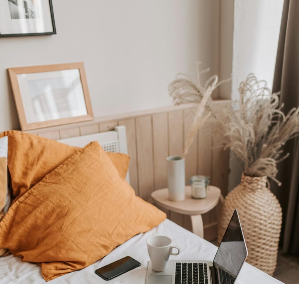 From above of open netbook with black screen and cup of coffee with smartphone and notebook placed on crumpled bed sheet in bedroom with big vase on floor